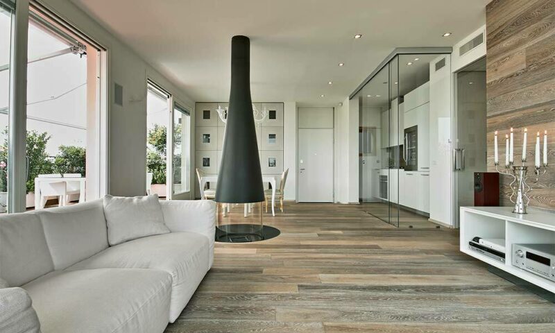 Modern living room with white furniture, hardwood floors, large windows, and a center fireplace.