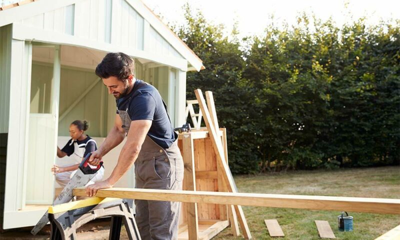 Carpenter cuts wood beam outside small building.