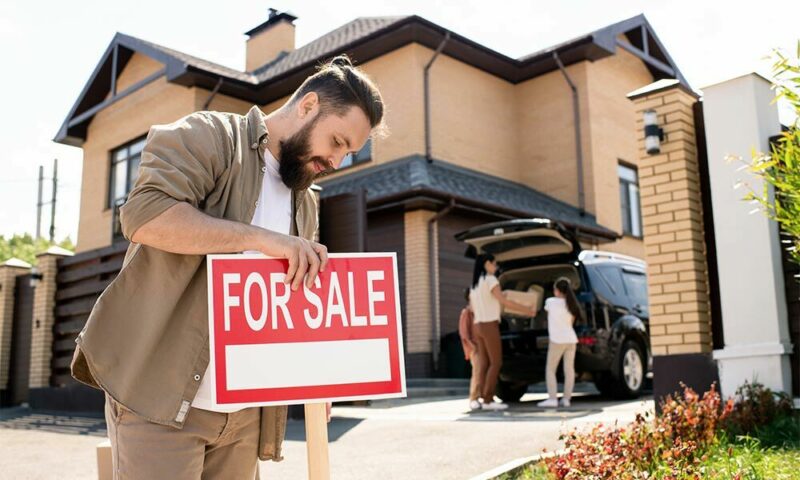 Man puts up a "for sale" sign while his family packs their car outside their home.