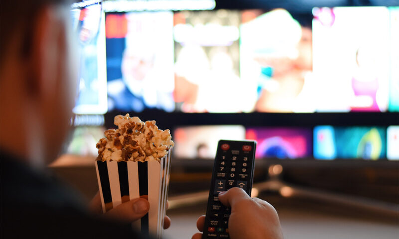 Over the shoulder view of man using remote to select a movie while holding popcorn.