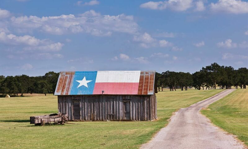 Old barn in a field with a Texas flag painted on the roof with trees in the background.