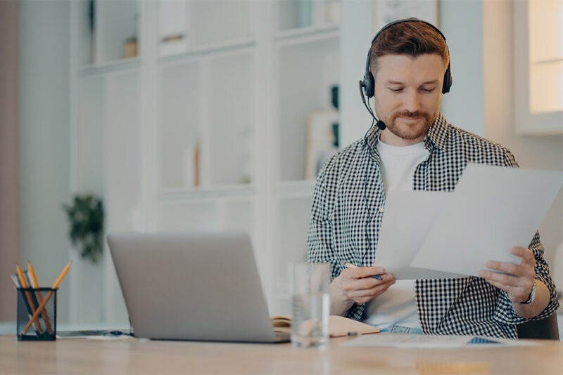 man smiling and shuffling through forms while working at his laptop