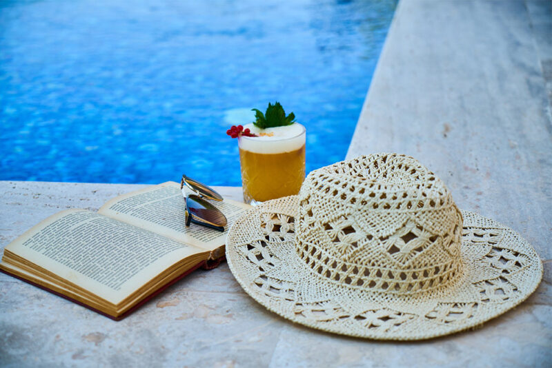 Sunhat, fancy and fruity cocktail, and book sitting next to an outdoor pool