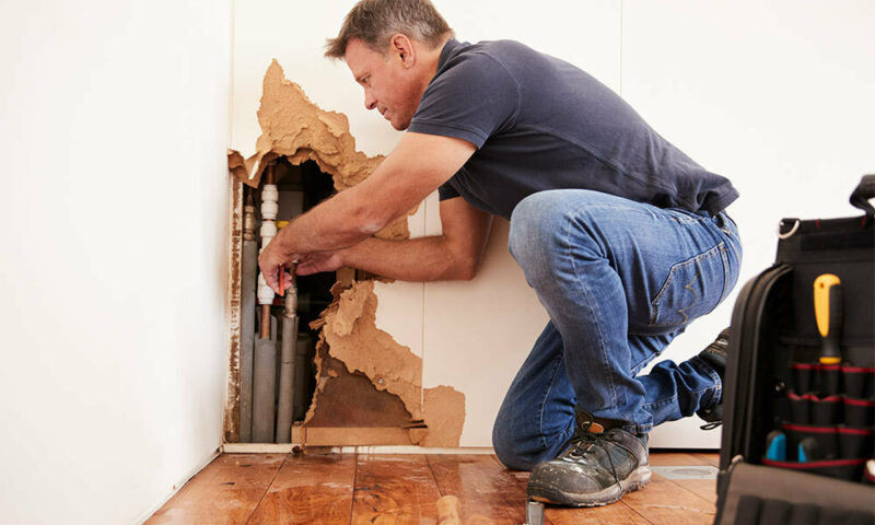 Man repairing busted pipes in corner of a room