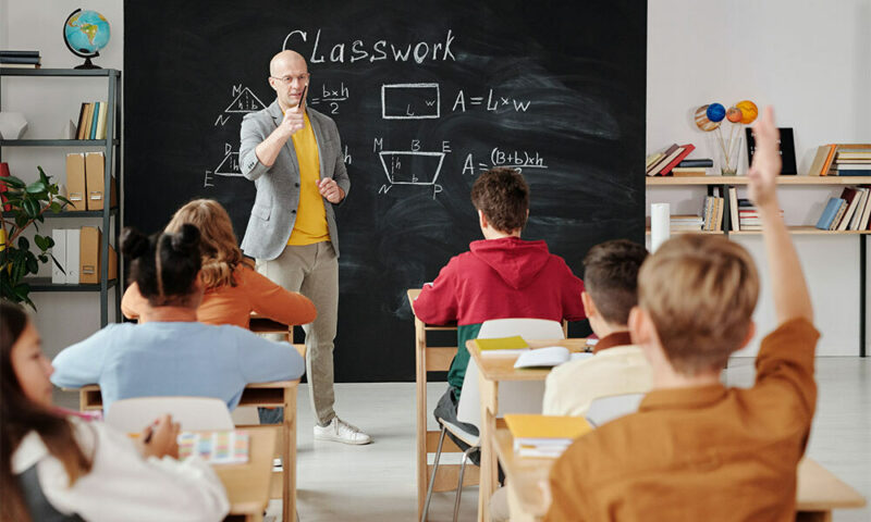 teacher in front of chalkboard in class asking students questions