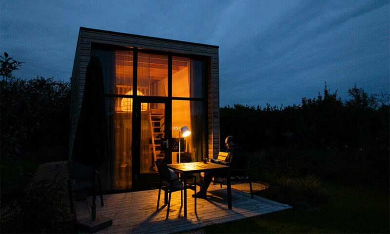 Man reading at dusk in front of tiny house with warm light coming from inside.